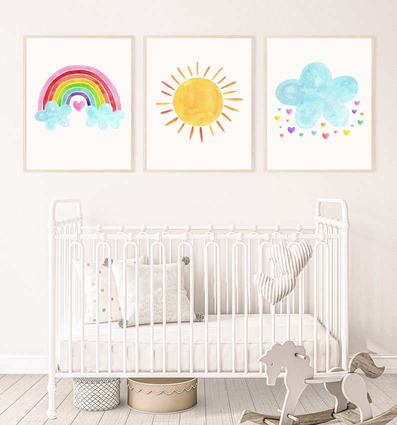 May include: Three watercolor prints featuring a rainbow with clouds, a sun, and a blue cloud with colorful hearts. The prints are displayed on a white wall above a white crib with a white mattress and two white pillows. There are two wooden rocking horses in the foreground.