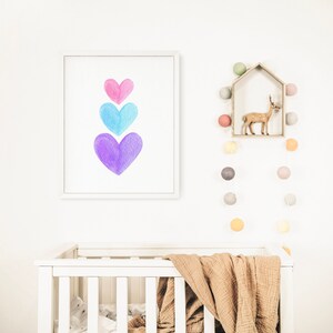 May include: A white crib with a brown blanket and a white frame. A framed print with three watercolor hearts in shades of pink, blue, and purple hangs on the wall above the crib. A string of colorful lights hangs on the wall to the right of the crib.