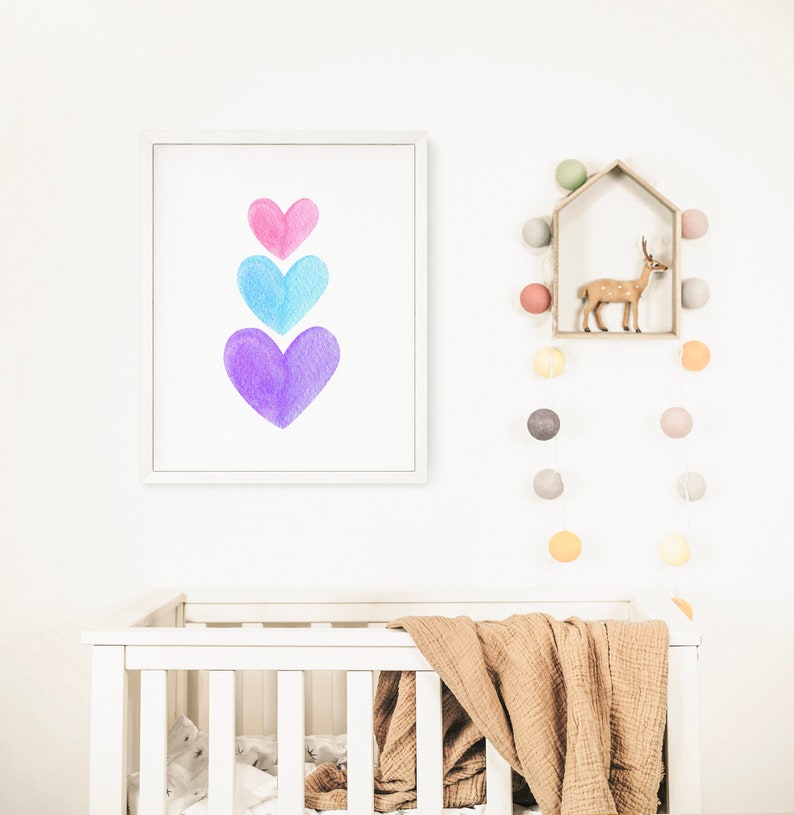 May include: A white crib with a brown blanket and a white frame. A framed print with three watercolor hearts in shades of pink, blue, and purple hangs on the wall above the crib. A string of colorful lights hangs on the wall to the right of the crib.