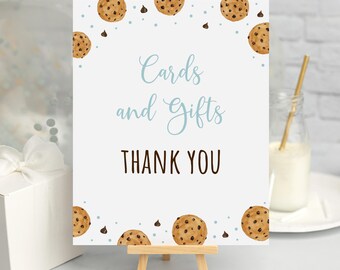 May include: A white sign with blue text that reads "Cards and Gifts Thank You". The sign is decorated with brown chocolate chip cookies and blue chocolate chips. The sign is on a small wooden easel.