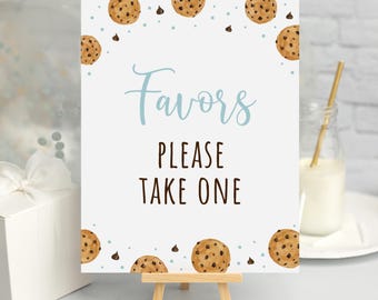 May include: A white sign with blue text that says "Favors Please Take One". The sign is decorated with brown chocolate chip cookies and blue chocolate chips. The sign is on a small wooden easel.