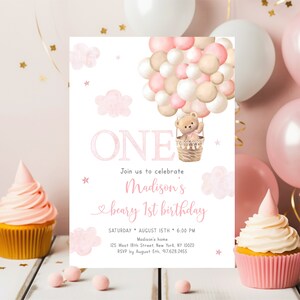 May include: Pink and beige balloon hot air balloon with a teddy bear in the basket. The balloon is surrounded by clouds and stars. The text "ONE" is in pink and the text "Join us to celebrate Madison's beary 1st birthday" is in pink and black. The text "SATURDAY AUGUST 15TH 6:00 PM" is in black. The text "Madison's home" is in pink. The text "125 West 18th Street, New York, NY 10020" is in black. The text "RSVP by August 5th. 917.628.2455" is in black. This is a first birthday party invitation.