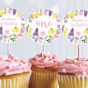 May include: Three round cupcake toppers with a floral design. The toppers feature a pink and yellow floral wreath with purple flowers and green leaves. The text on the toppers reads "Madison's 1st Birthday" and "one".