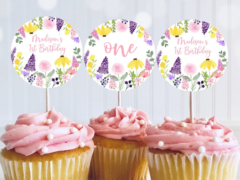 May include: Three round cupcake toppers with a floral design. The toppers feature a pink and yellow floral wreath with purple flowers and green leaves. The text on the toppers reads "Madison's 1st Birthday" and "one".