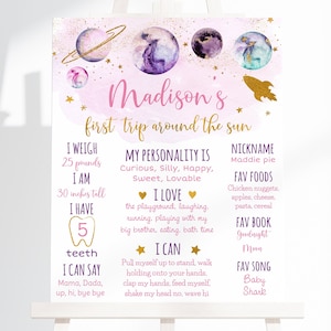 May include: A pink and purple space-themed first birthday sign with the name "Madison's" and the text "first trip around the sun". The sign lists details about the child, including their weight, height, favorite foods, and songs.