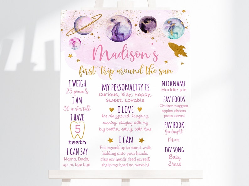 May include: A pink and purple space-themed first birthday sign with the name "Madison's" and the text "first trip around the sun". The sign lists details about the child, including their weight, height, favorite foods, and songs.