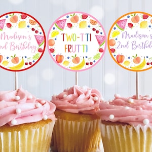 May include: Three cupcakes with pink icing and white sprinkles. Each cupcake has a circular topper with a fruit design and text. The toppers read "Madison's 2nd Birthday" and "TWO-TTI FRUTTI". The toppers have red, pink, and orange borders.