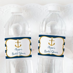 K&ouml;nnte beinhalten: Zwei transparente Plastikwasserflaschen mit wei&szlig;en Deckeln. Jede Flasche hat ein marineblau-wei&szlig; gestreiftes Etikett mit einem goldenen Anker und dem Text "Alyssa's Bridal Shower". Die Etiketten haben einen goldenen Rand.