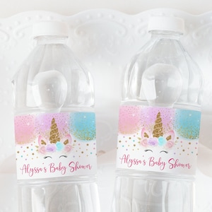 Può includere: Due bottiglie d'acqua in plastica trasparente con etichette rosa, blu e dorate che mostrano un'illustrazione di unicorno e il testo "Alyssa's Baby Shower".