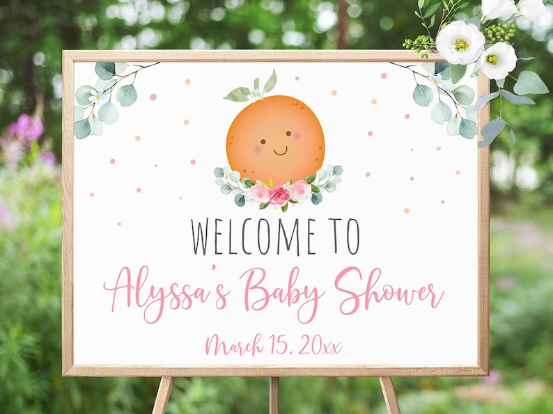 May include: A white welcome sign with a pink and green floral design and a smiling orange illustration. The text reads "Welcome to Alyssa's Baby Shower March 15. 20xx".
