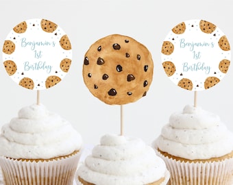May include: Three cupcake toppers featuring a chocolate chip cookie design. The toppers are white with blue text that reads "Benjamin's 1st Birthday".