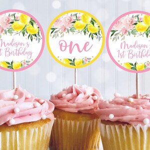 May include: Three pink and yellow cupcake toppers with lemon and floral designs. The toppers say "Madison's 1st Birthday" and "one".