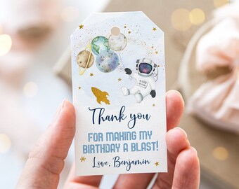May include: A blue and white thank you tag with a watercolor illustration of an astronaut holding balloons shaped like planets. The tag has gold glitter and stars and the text "Thank you for making my birthday a blast! Love, Benjamin".