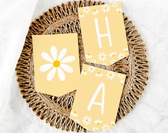 Peut inclure: Une bannière jaune avec des marguerites blanches et les lettres "HA" en blanc. La bannière est sur un dessous de plat en osier brun.