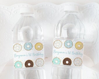 May include: Two clear plastic water bottles with personalized labels featuring a donut theme. The labels are white with blue and yellow sprinkles and feature various donuts with different toppings. The text on the labels reads "Benjamin's 1st Birthday".