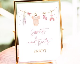 Op de afbeelding: Een printbaar bord met een roze en gouden kleurschema. Het bord heeft een string met baby-thema decoraties, waaronder een rompertje, schoentjes, een rammelaar en een regenboog. De tekst op het bord luidt "Sweets and Treats Enjoy!"