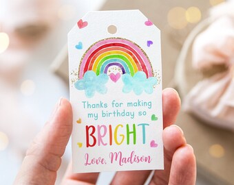 May include: A white gift tag with a rainbow design, blue clouds, and gold glitter accents. The tag reads "Thanks for making my birthday so BRIGHT. Love, Madison" in colorful lettering. Small hearts and stars decorate the tag.