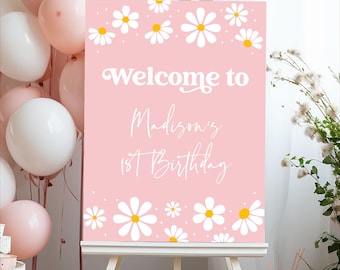 May include: A pink sign on an easel with the text "Welcome to Madison's 1st Birthday". The sign is decorated with white daisies with yellow centers. Balloons in pink and white are in the background.