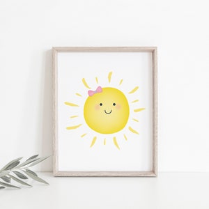 Puede incluir: Una impresi&oacute;n enmarcada de un sol amarillo con un lazo rosa, sonriendo con mejillas sonrosadas. El sol tiene rayos amarillos que irradian de &eacute;l.