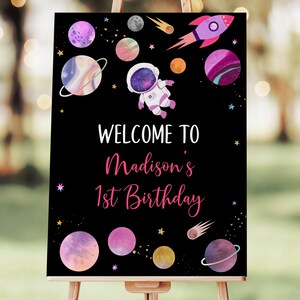 May include: A black welcome sign with a pink and white astronaut, planets, and stars. The sign says "Welcome to Madison's 1st Birthday".