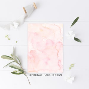 May include: A rectangular card with a watercolor design in shades of pink and peach, with the text "OPTIONAL BACK DESIGN". The card is surrounded by white flowers, green leaves, and a spool of ribbon on a white wooden surface.