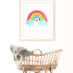 May include: A framed watercolor painting of a rainbow with clouds and a pink heart in the center. The painting is hanging on a white wall above a wicker bassinet.