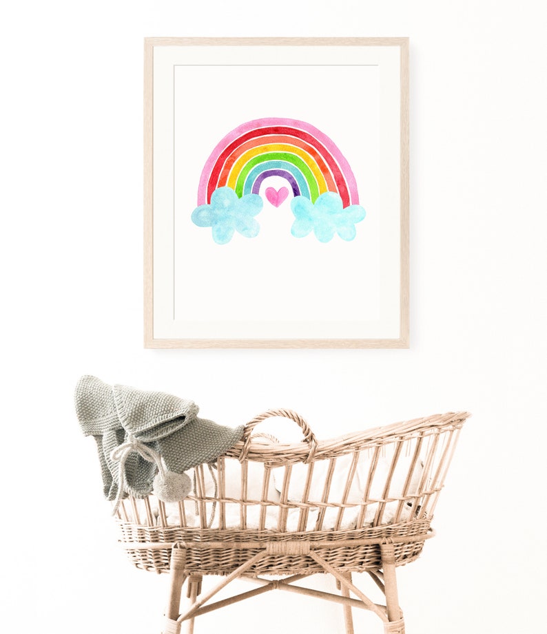 May include: A framed watercolor painting of a rainbow with clouds and a pink heart in the center. The painting is hanging on a white wall above a wicker bassinet.