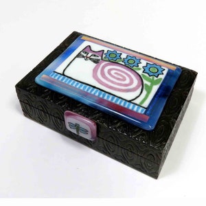 May include: A small wooden box with a black swirl pattern. The lid features a colorful glass inlay of a white cat with pink and blue swirls, surrounded by blue flowers. A dragonfly is on the latch.