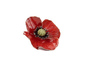 May include: A red poppy flower brooch with a black center and green stamen. The poppy is made of enamel and has a shiny finish.
