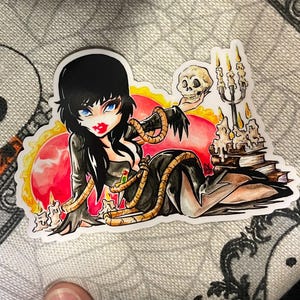 May include: A sticker featuring a cartoon illustration of a woman with black hair, blue eyes, and red lipstick. She is wearing a black dress and holding a skull. The background includes candles, books, and a red and yellow design.