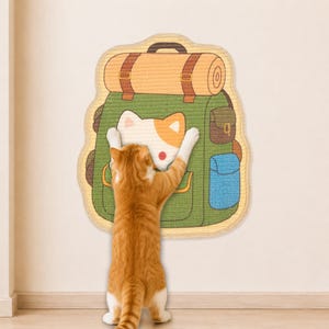 Könnte beinhalten: Ein an der Wand montiertes Kratzbrett für Katzen in Form eines grünen Wanderrucksacks mit einem beigen Schlafsack oben. Der Rucksack zeigt ein weiß-oranges Katzengesicht, eine kleine braune Seitentasche und eine blaue Seitentasche. Eine rote Katze.