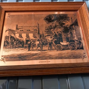 May include: Framed sepia-toned print depicting a horse-drawn carriage and figures in a town square. The print, titled "AFTERNOON STOP," is set in a wooden frame. The scene includes horses, buildings, and people, evoking a historical setting.
