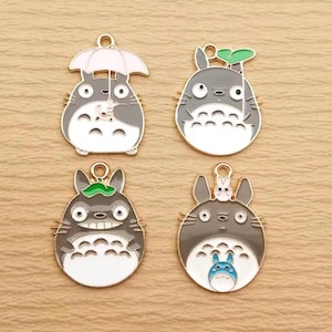 May include: Four enamel charms featuring the character Totoro from the animated film My Neighbor Totoro. Each charm is gray, white, and gold, with different designs including an umbrella, leaf, and small Totoro.