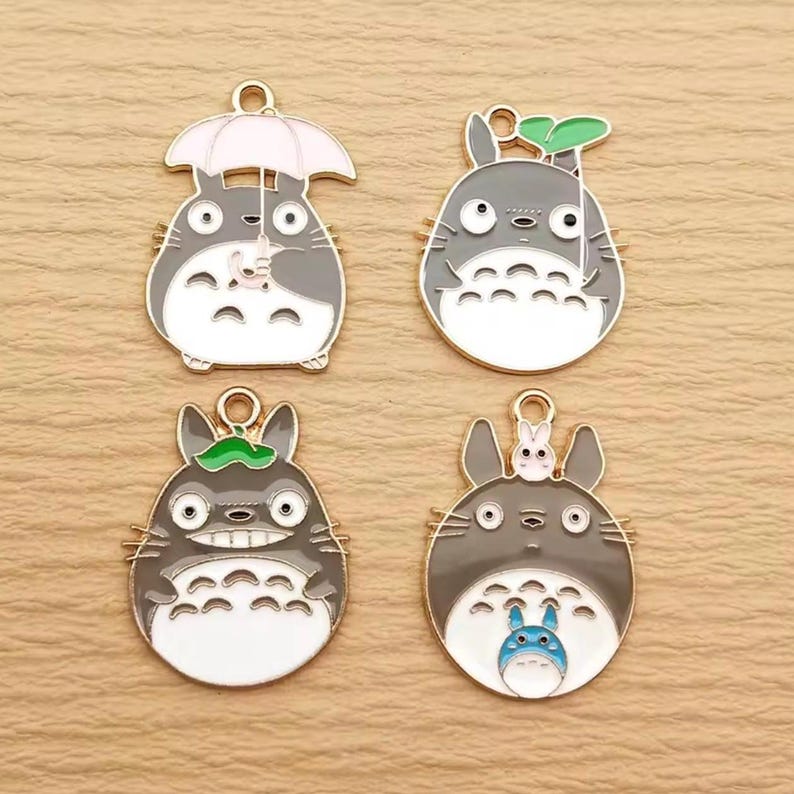 May include: Four enamel charms featuring the character Totoro from the animated film My Neighbor Totoro. Each charm is gray, white, and gold, with different designs including an umbrella, leaf, and small Totoro.