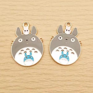 May include: Two gray and white enamel charms featuring a cartoon character with a small bunny on top. The charms have a gold-colored loop at the top for attaching to jewelry or other crafts. The character has a blue and white belly.