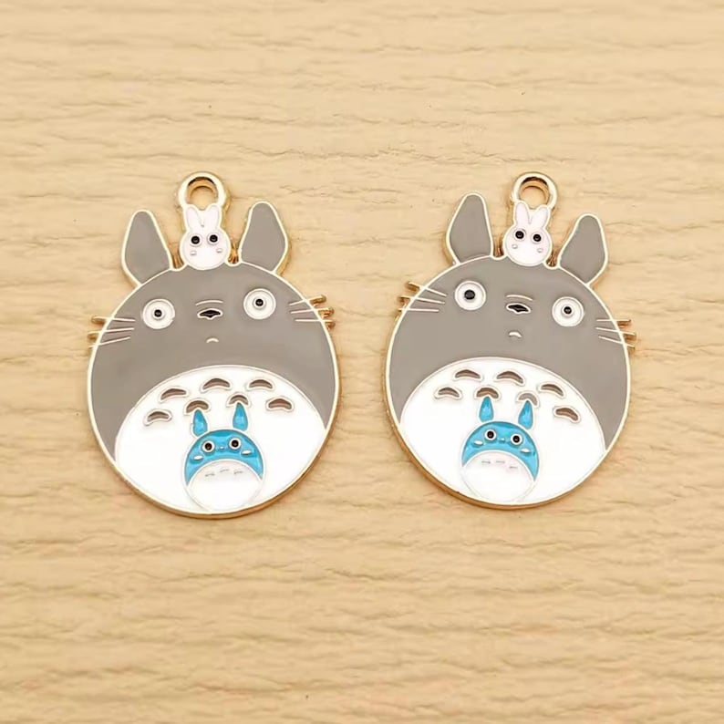 May include: Two gray and white enamel charms featuring a cartoon character with a small bunny on top. The charms have a gold-colored loop at the top for attaching to jewelry or other crafts. The character has a blue and white belly.
