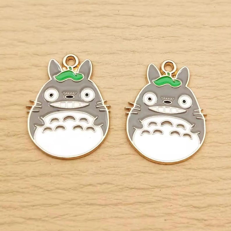 May include: Two gray and white enamel charms featuring a smiling cartoon character with a green leaf on its head. The charms have a gold-colored border and a loop at the top for attaching to jewelry or other crafts.