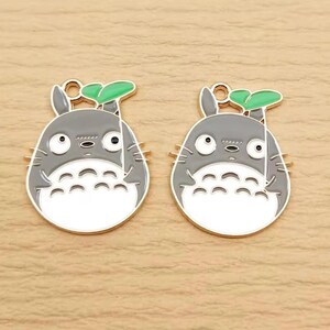 May include: Two gray and white enamel charms in the shape of a cartoon character, each with a green leaf and gold-colored accents. The charms have a loop at the top for attaching to jewelry or other crafts. The design is a popular anime character.