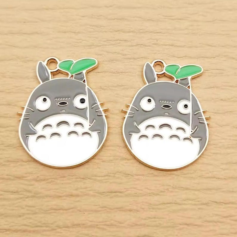 May include: Two gray and white enamel charms in the shape of a cartoon character, each with a green leaf and gold-colored accents. The charms have a loop at the top for attaching to jewelry or other crafts. The design is a popular anime character.