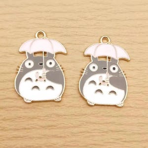May include: Two enamel charms featuring a cartoon character with a gray body, white belly, and a white umbrella. The charms have a gold-colored loop at the top for attaching to jewelry or other crafts. The character is holding the umbrella.