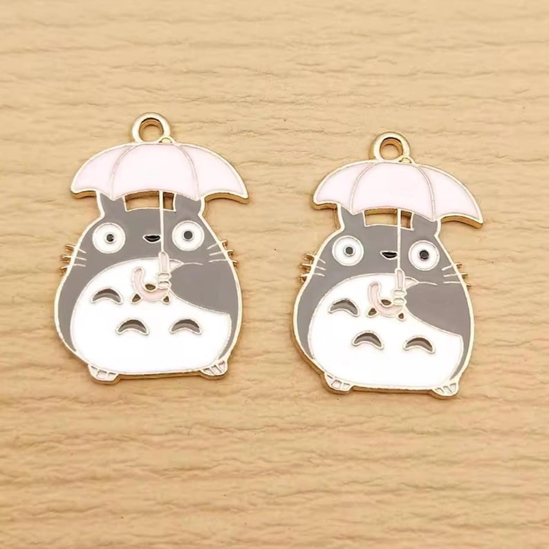 May include: Two enamel charms featuring a cartoon character with a gray body, white belly, and a white umbrella. The charms have a gold-colored loop at the top for attaching to jewelry or other crafts. The character is holding the umbrella.