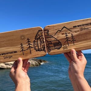 Può includere: Due taglieri in legno tenuti contro un cielo blu e il mare. Ogni tagliere presenta un design unico: un camper, montagne, alberi, sole e testo in tedesco. I taglieri sono incisi con la frase "DER WEG IST DAS ZIEL" e nomi.