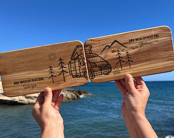 Könnte beinhalten: Zwei Holzschneidebretter vor blauem Himmel und Meer. Jedes Brett hat ein einzigartiges Design: ein Wohnmobil, Berge, Bäume, Sonne und deutscher Text. Die Bretter sind mit dem Satz "DER WEG IST DAS ZIEL" und Namen graviert.