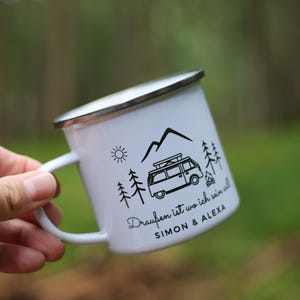 May include: White enamel camping mug with a black and white design of a van, mountains, trees, and a campfire. The text on the mug reads "Drau&szlig;en ist wo ich sein will SIMON & ALEXA".