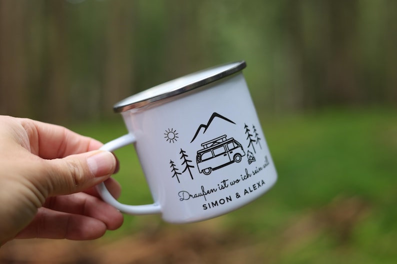 May include: White enamel camping mug with a black and white design of a van, mountains, trees, and a campfire. The text on the mug reads "Drau&szlig;en ist wo ich sein will SIMON & ALEXA".