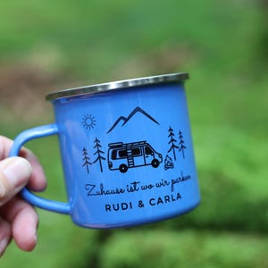 May include: Blue enamel camping mug with a black and white illustration of a camper van, mountains, and trees. The mug has the text "Zuhause ist wo wir parken" and "RUDI & CARLA" printed on it.