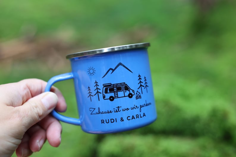May include: Blue enamel camping mug with a black and white illustration of a camper van, mountains, and trees. The mug has the text "Zuhause ist wo wir parken" and "RUDI & CARLA" printed on it.