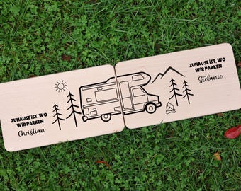 Könnte beinhalten: Zwei Holzschilder mit einem Wohnmobil-Design, Bäumen, Sonne und Bergen. Der Text "Zuhause ist, wo wir parken Christian" und "Zuhause ist, wo wir parken Stefanie" ist auf den Schildern aufgedruckt. Die Schilder liegen auf einer grünen, grasbewachsenen Oberfläche.