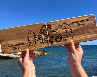 Könnte beinhalten: Zwei Holzschneidebretter vor blauem Himmel und Meer. Jedes Brett hat ein einzigartiges Design: ein Wohnmobil, Berge, Bäume, Sonne und deutscher Text. Die Bretter sind mit dem Satz "DER WEG IST DAS ZIEL" und Namen graviert.
