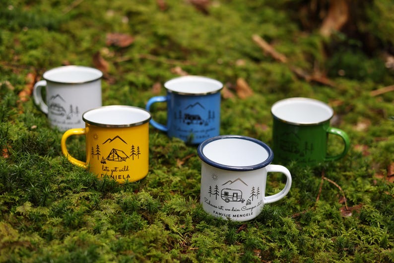 May include: Five enamel camping mugs with different designs and colours. The mugs are on a bed of green moss. The designs include a tent, a camper, and mountains. The text on the mugs reads "Let's get wild Daniela" and "Adventure is my favourite campground Family Mengel".